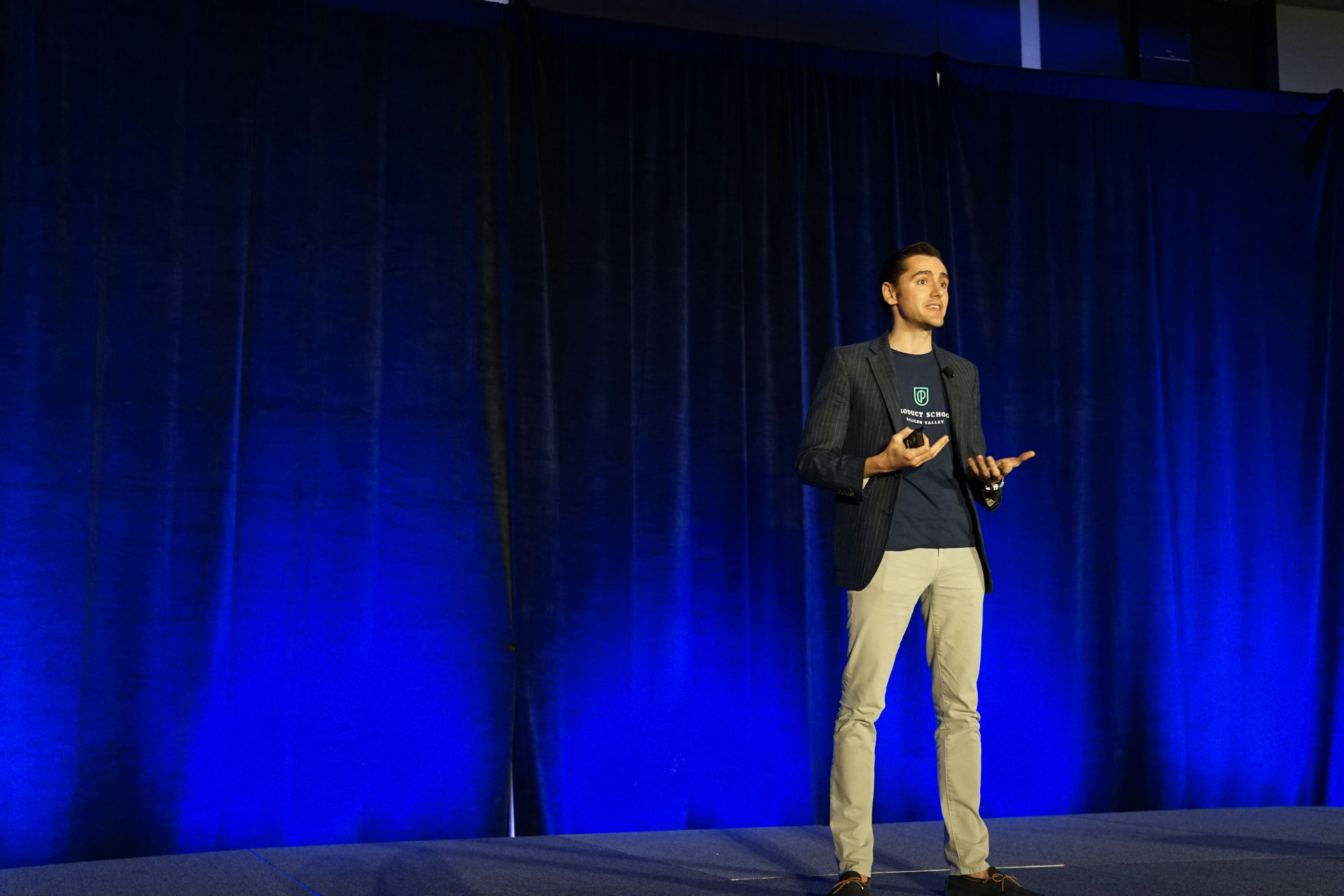 Photo of a man giving a talk on stage with blue curtains behind him.